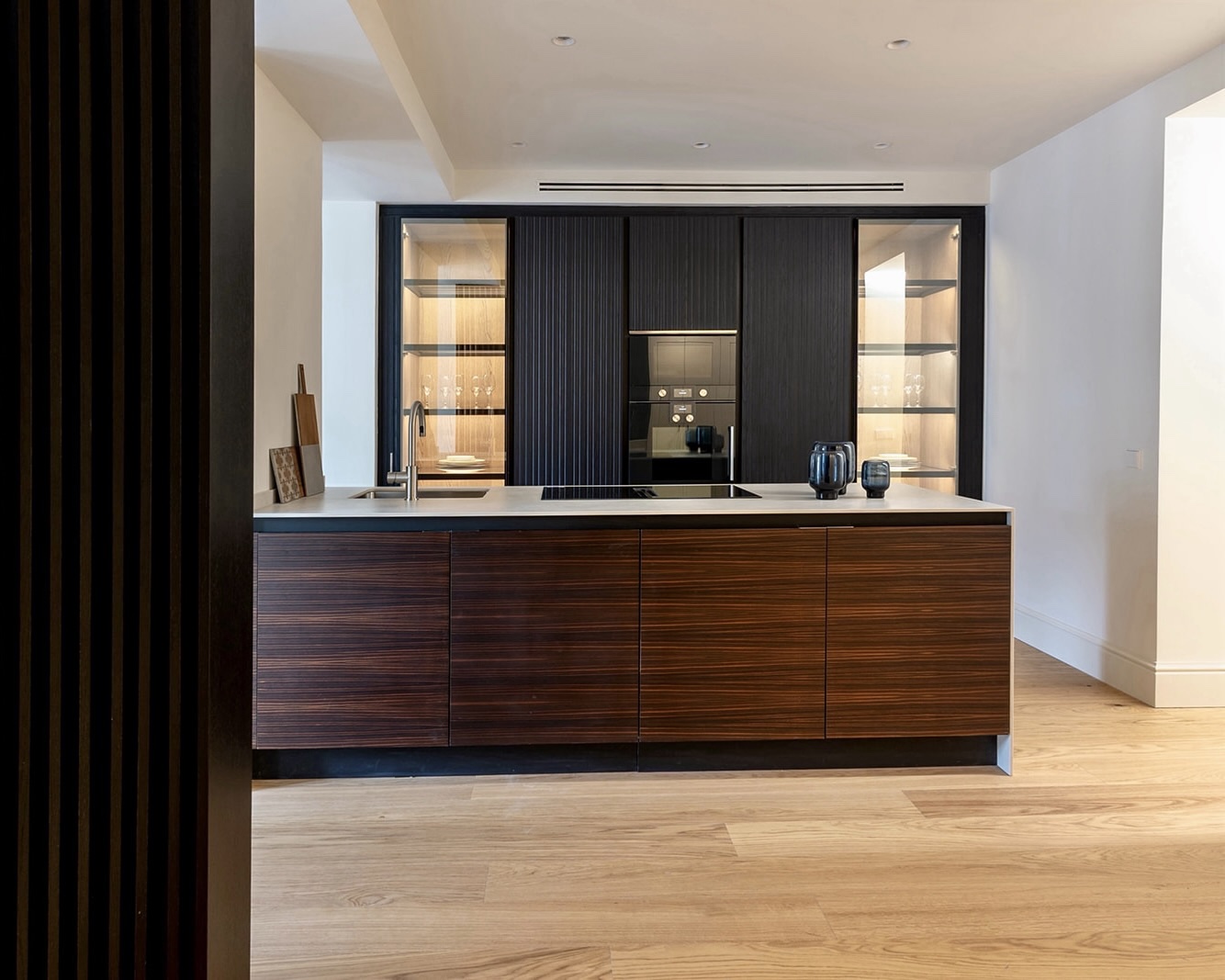 Cocina moderna con isla de madera oscura, armarios negros y estantes iluminados. Electrodomésticos integrados y encimera blanca. Diseño minimalista con acabados de alta gama en un espacio luminoso con suelo de madera clara.
