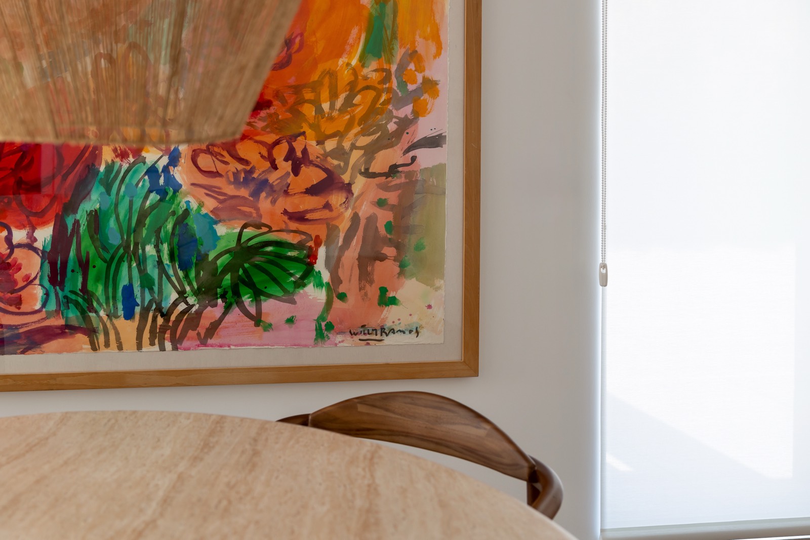 Pintura abstracta colorida en un marco de madera colgada en una pared blanca. Se observa parte de una mesa redonda en primer plano y una ventana con persiana enrollable al lado.