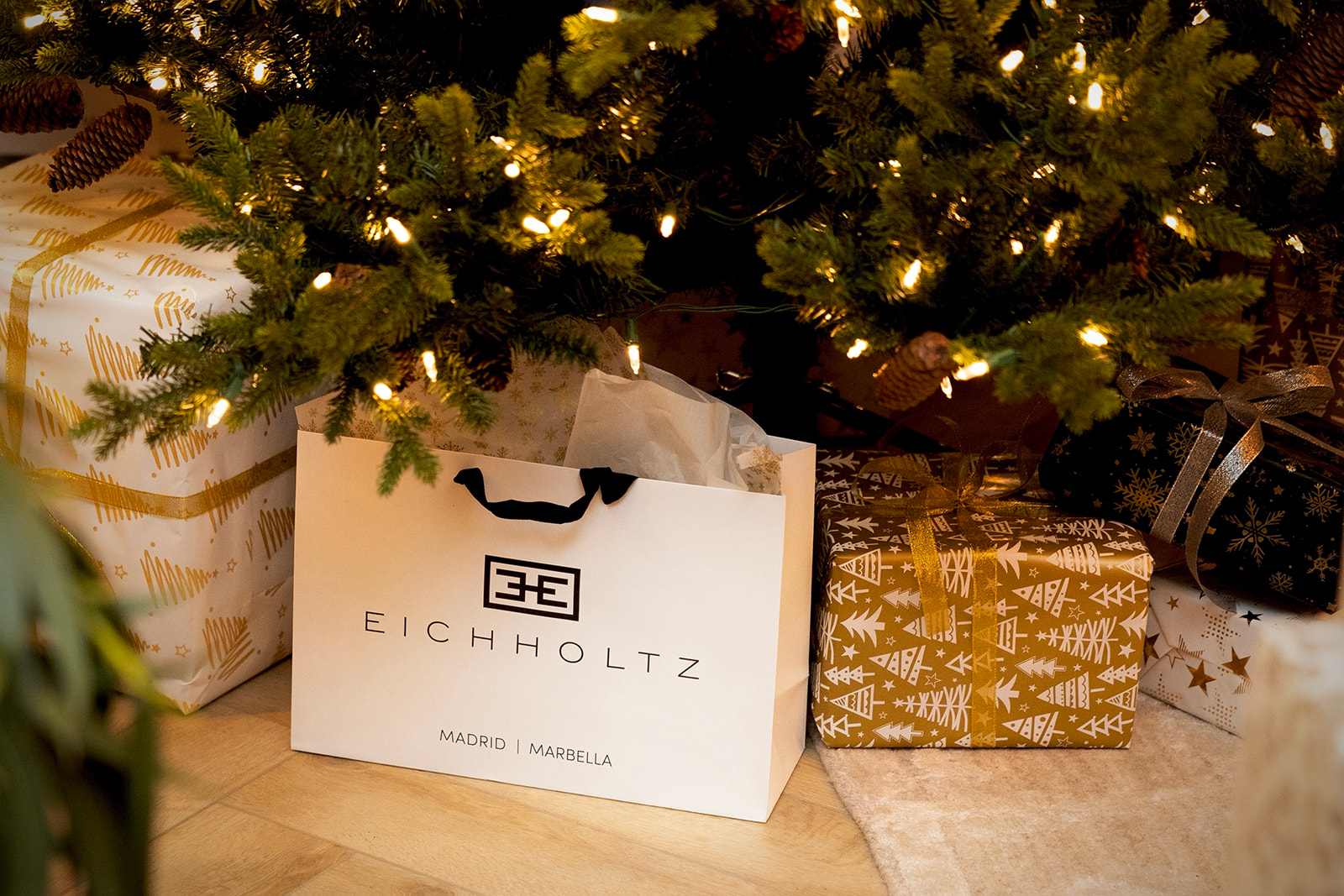 Bolsa de compras de Eichholtz junto a regalos envueltos bajo un árbol de Navidad iluminado con luces cálidas. La escena muestra una atmósfera festiva y acogedora.