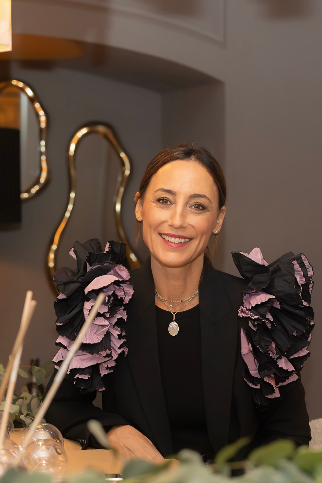 Mujer sonriente con chaqueta negra adornada con volantes negros y rosados en los hombros. Lleva un collar con colgante y está sentada en un entorno elegante con decoración dorada visible en el fondo.