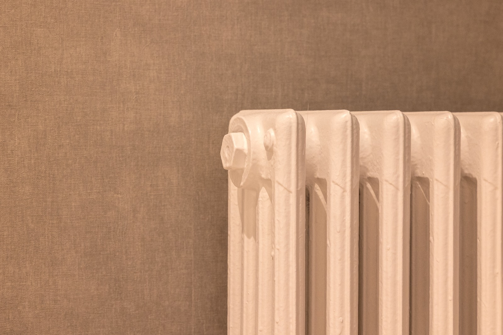 Primer plano de un radiador clásico de color beige con columnas verticales contra una pared de tono marrón claro. La imagen tiene un aspecto monocromático y cálido.