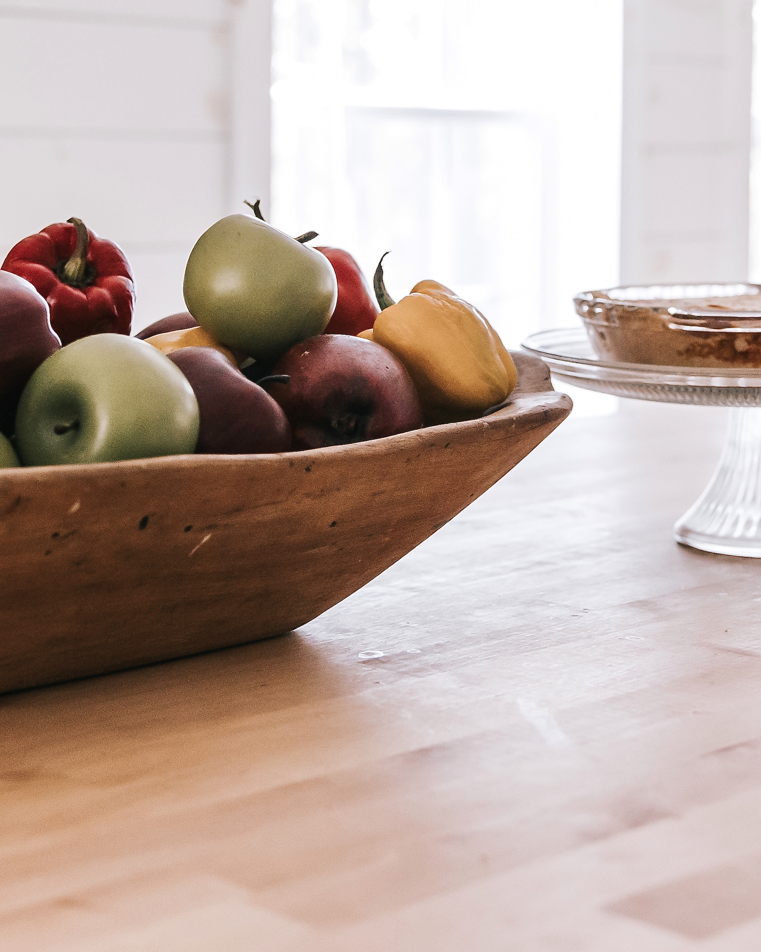 Frutero rústico de madera con manzanas verdes y rojas junto a pimientos, sobre una mesa clara. En el fondo se aprecia un recipiente de cristal