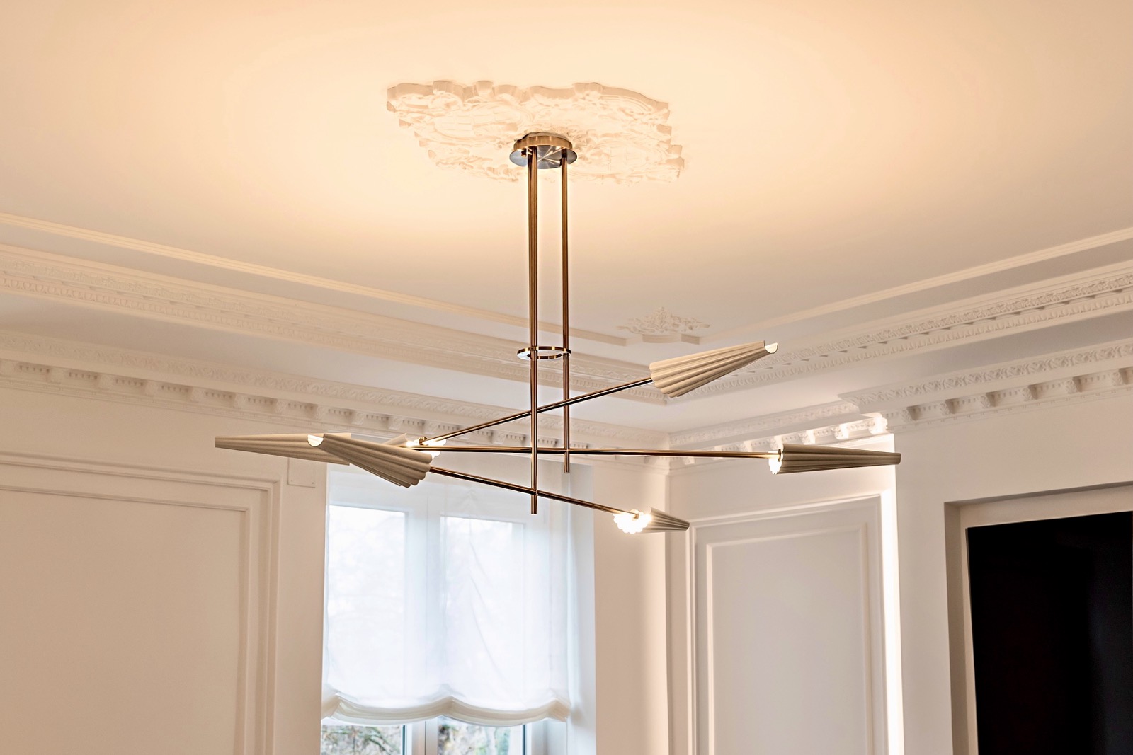 Lámpara de techo moderna con brazos metálicos y luces LED, colgando de un techo blanco ornamentado con molduras clásicas en una habitación elegante