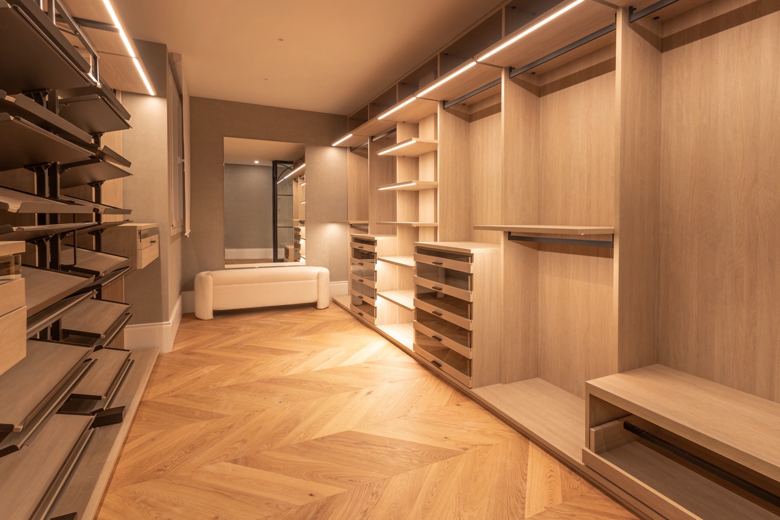 Amplio vestidor moderno con muebles de madera clara, iluminación LED empotrada, suelo de parquet en espiga y un sofá blanco. El diseño incluye estanterías, cajones y barras para colgar ropa, creando un espacio organizado y elegante.