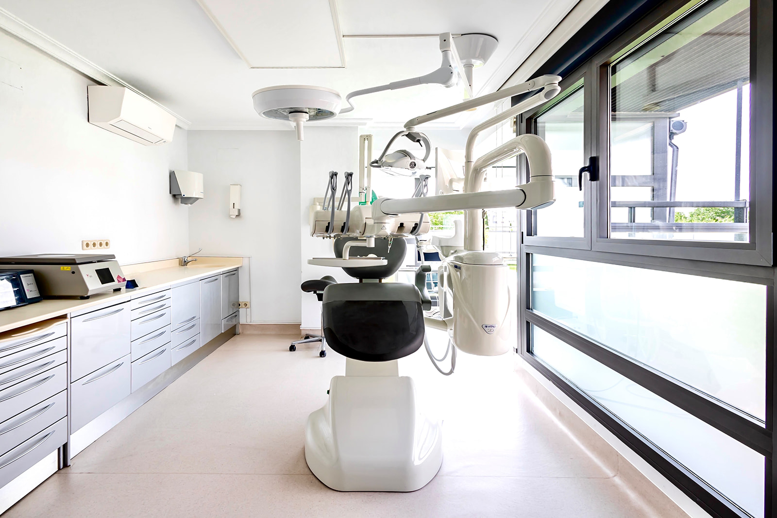 Consultorio dental moderno y luminoso con sillón dental blanco, equipos médicos, armarios de almacenamiento y ventanas grandes que permiten la entrada de luz natural