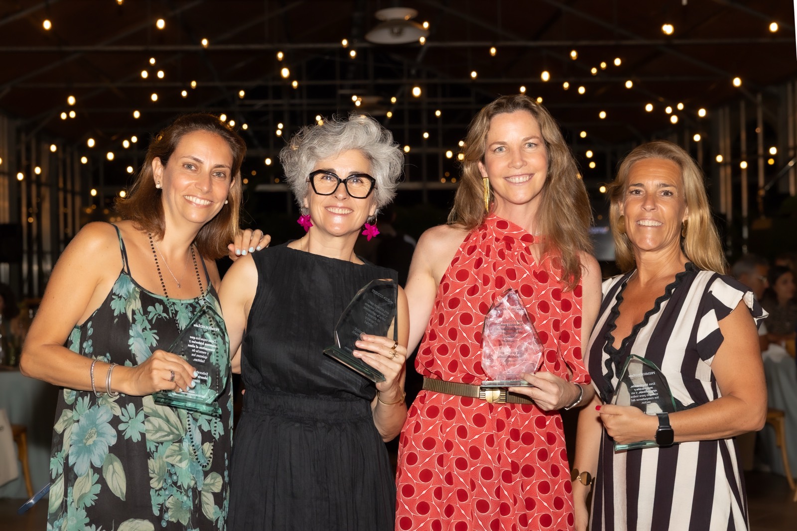 Cuatro mujeres sonrientes sosteniendo premios bajo luces decorativas, vestidas con diferentes estilos y colores