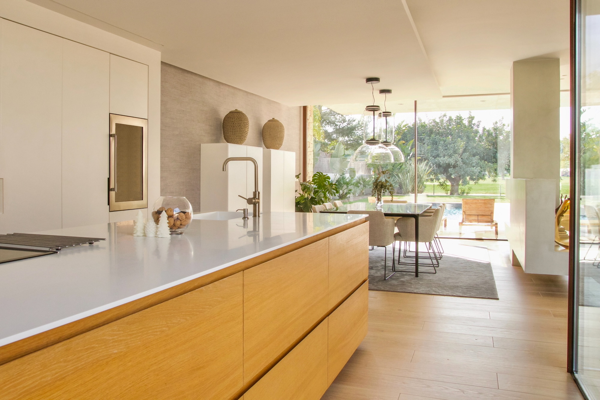 Cocina moderna con isla de madera clara y encimera blanca, decorada con jarrones artesanales. Espacio abierto que conecta con comedor y jardín a través de ventanales. Diseño minimalista con acabados limpios y luminosos.