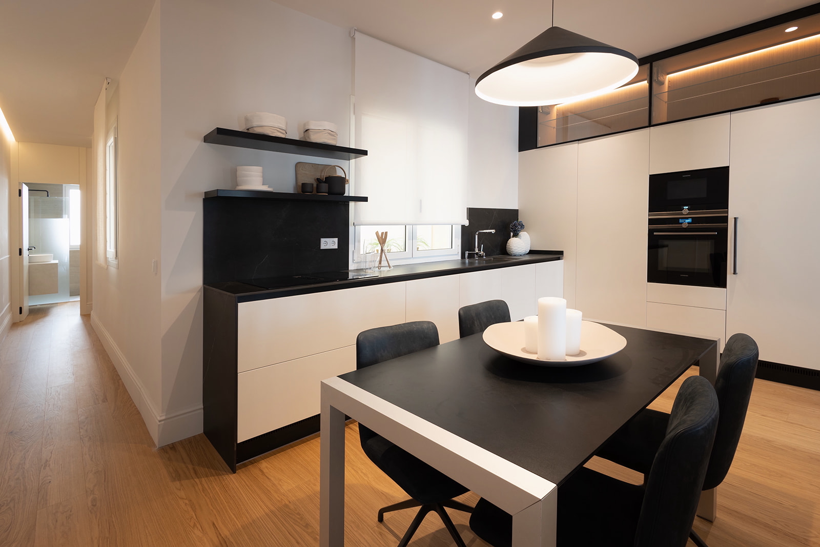 Cocina moderna con diseño minimalista en blanco y negro. Muebles blancos, encimera negra, mesa de comedor negra con sillas a juego. Lámpara colgante grande y estantes negros con accesorios blancos. Piso de madera y electrodomésticos integrados.