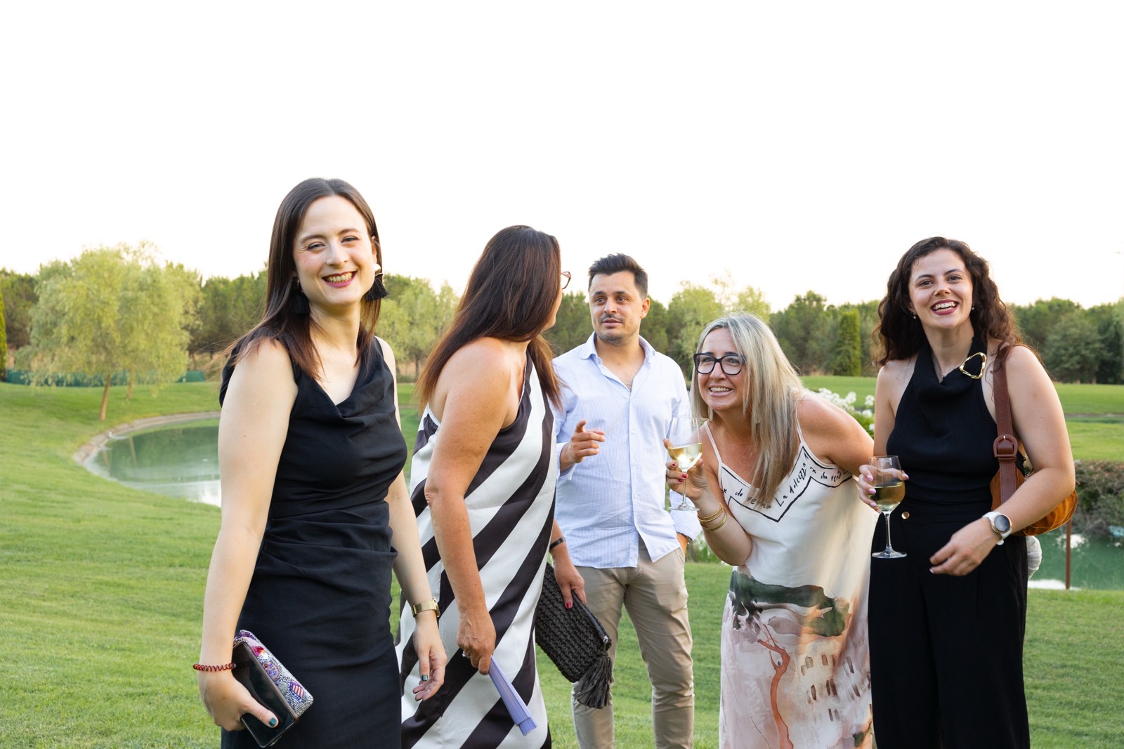 Grupo de cinco personas elegantemente vestidas sonriendo y sosteniendo copas de vino en un jardín con un lago en el fondo