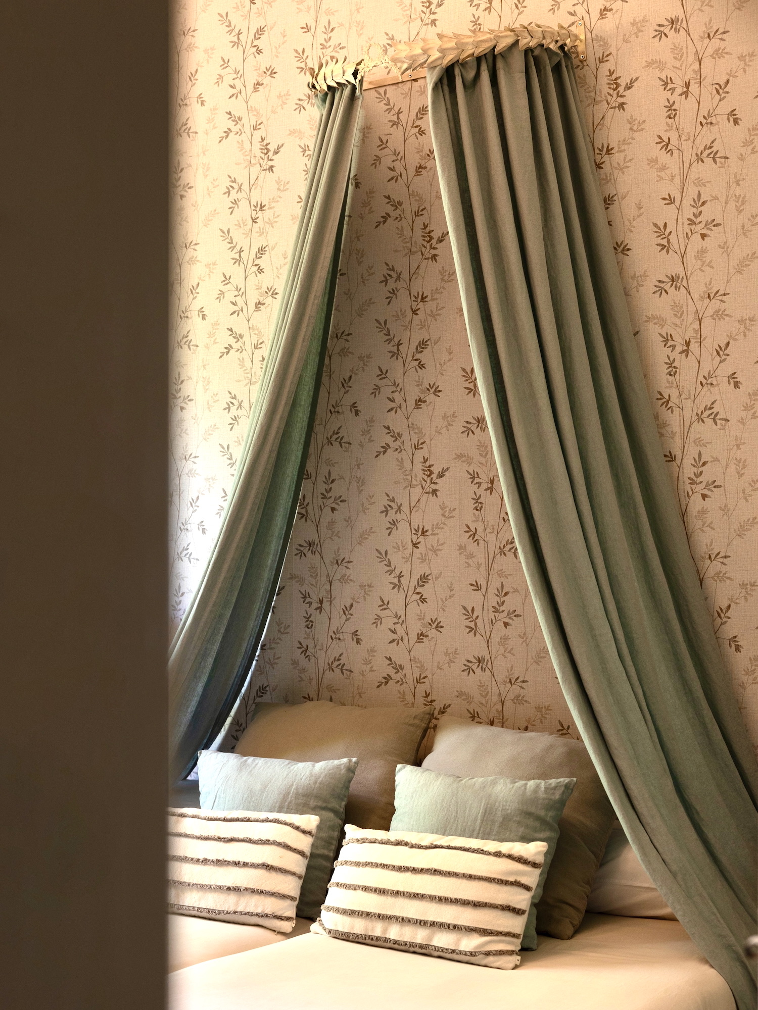 Elegante dosel de tela verde sage sobre una cama con almohadas decorativas rayadas, contra una pared con papel tapiz floral en tonos beige. El dosel crea un ambiente acogedor y romántico en el dormitorio.