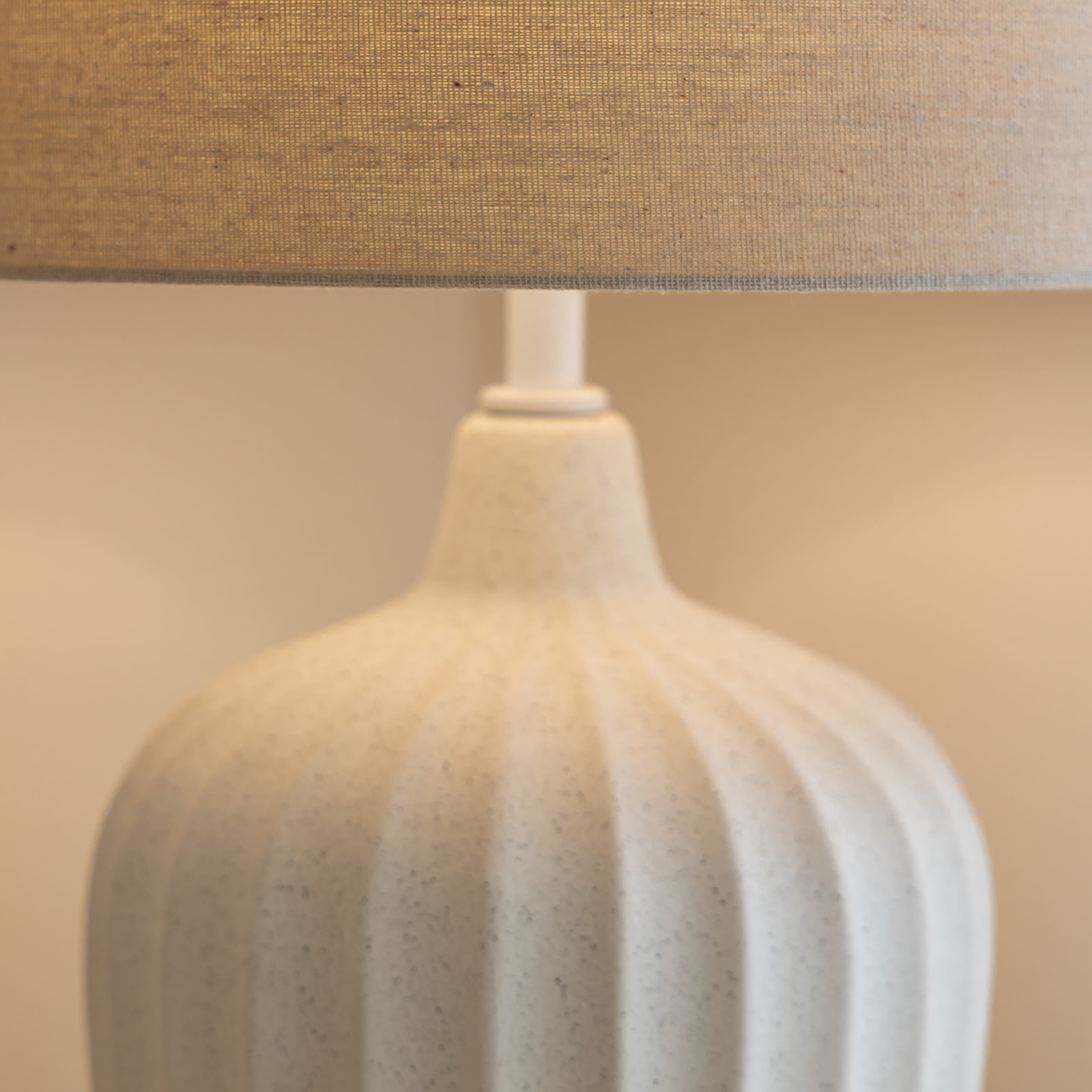 Detalle de una lámpara de mesa con base de cerámica en color beige claro con forma acanalada y pantalla cilíndrica de tela en tono marrón claro.