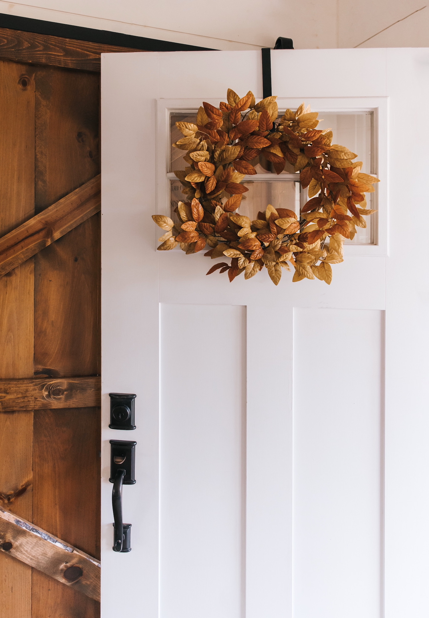 Corona decorativa de hojas doradas y marrones colgada en una puerta blanca con detalles en madera. La puerta tiene un diseño craftsman con paneles y herrajes negros tradicionales