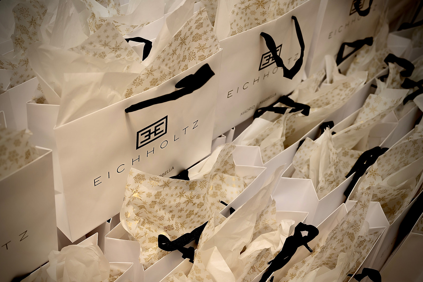 Varias bolsas de compra blancas con el logotipo de Eichholtz, rodeadas de papel de seda decorativo con copos de nieve dorados. Las bolsas están apiladas y mezcladas con el papel de seda, creando una imagen elegante y festiva.
