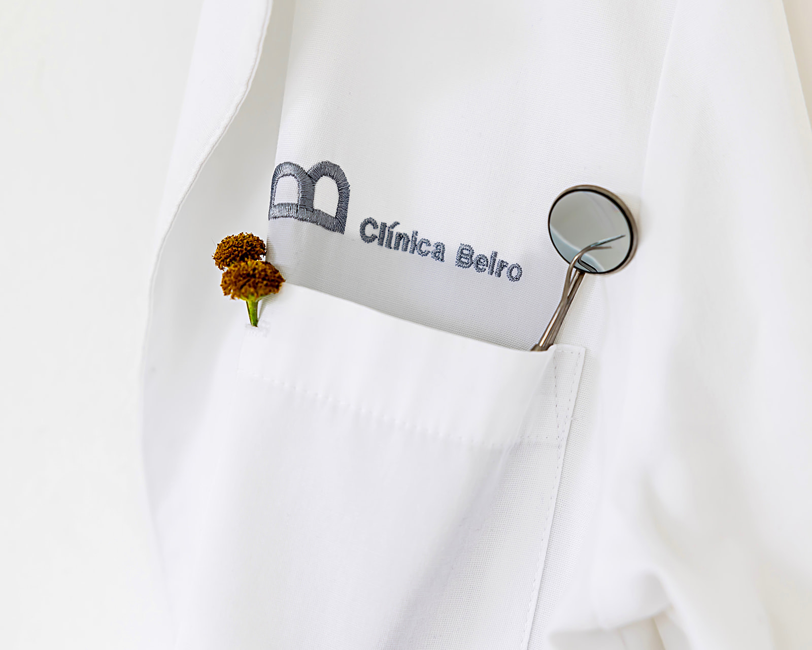 Bata médica blanca con el logo bordado de Clínica Beiro. En el bolsillo se observa un espejo dental y una flor pequeña de color marrón.