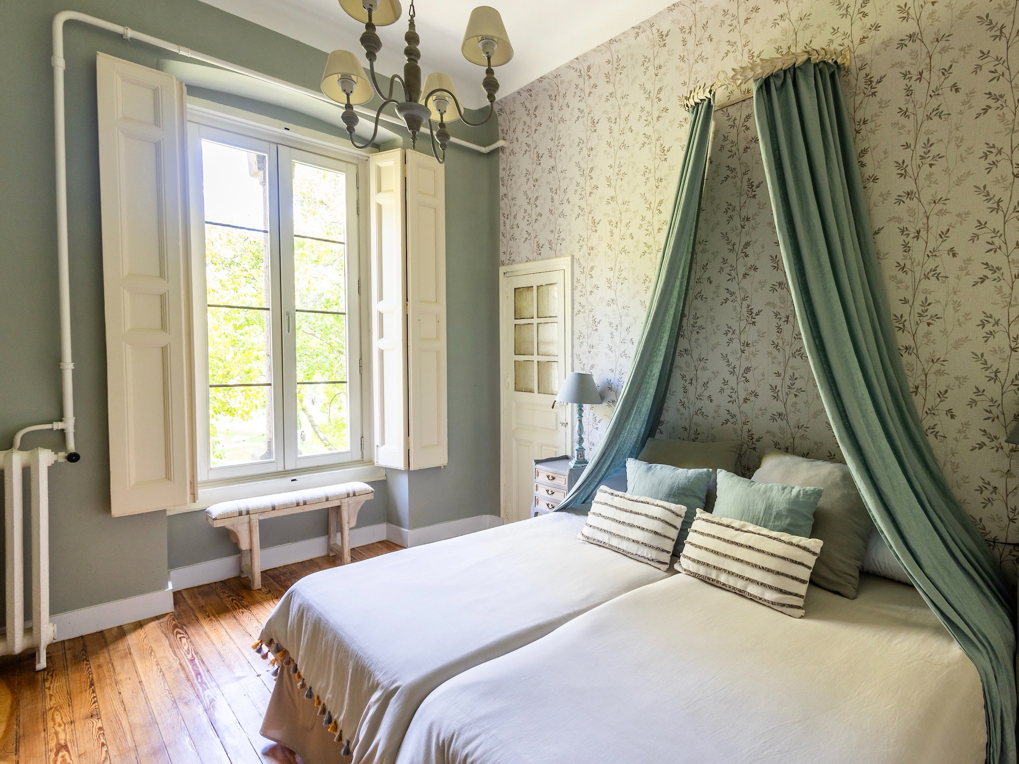 Dormitorio elegante con paredes en verde sage y papel tapiz floral, cama con dosel verde, ventana con contraventanas blancas, piso de madera y lámpara de araña clásica. Decoración que combina elementos vintage y modernos.