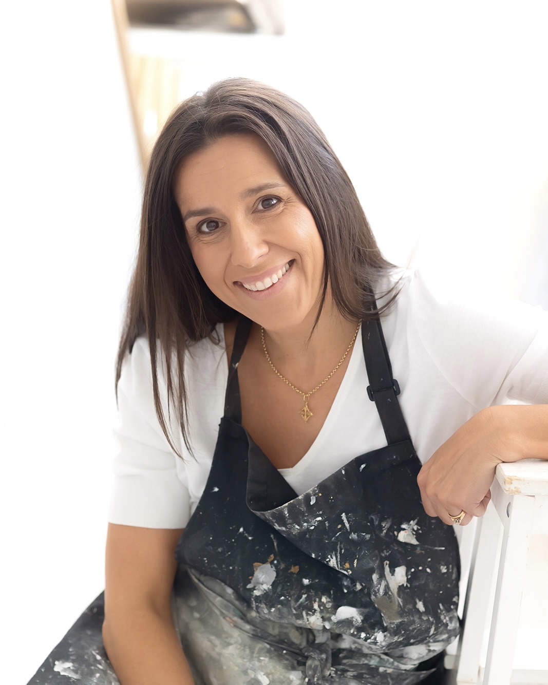 Mujer sonriente con cabello largo y oscuro vistiendo una camisa blanca y un delantal negro manchado de pintura. Lleva un collar dorado y parece estar en un ambiente interior luminoso, posiblemente un estudio de arte o taller.