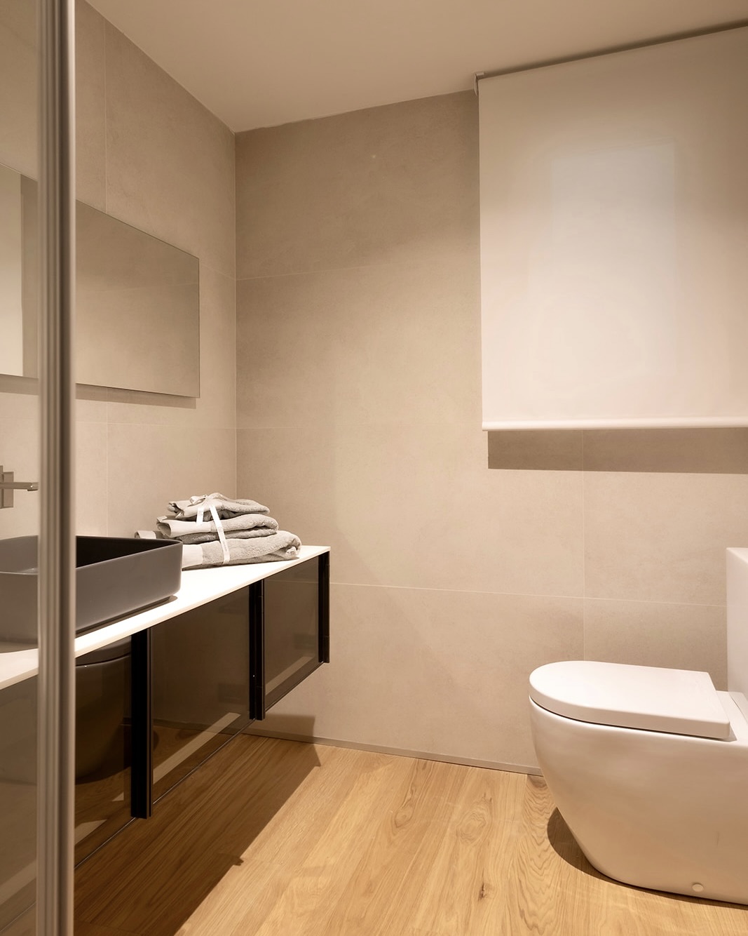 Baño moderno y minimalista con paredes beige, suelo de madera, lavabo negro sobre mueble oscuro, espejo rectangular y sanitario blanco. Toallas dobladas sobre el lavabo y una cortina enrollable en la ventana.