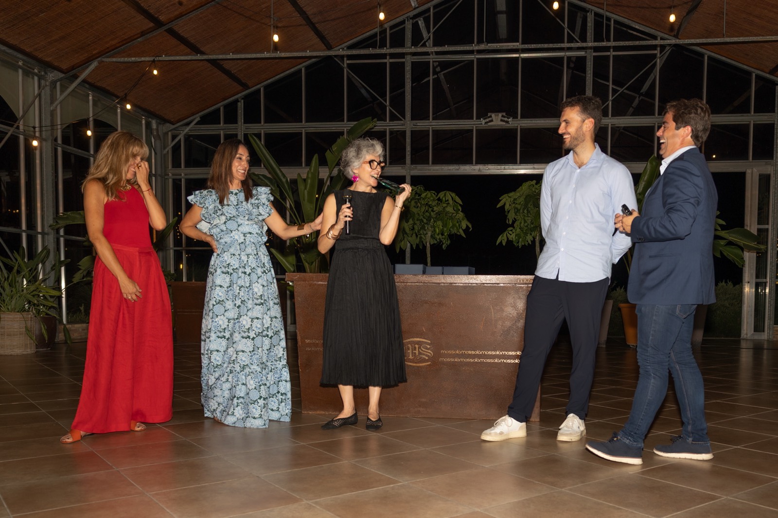 Grupo de cinco personas vestidas elegantemente conversando en un espacio interior con iluminación cálida y plantas de fondo