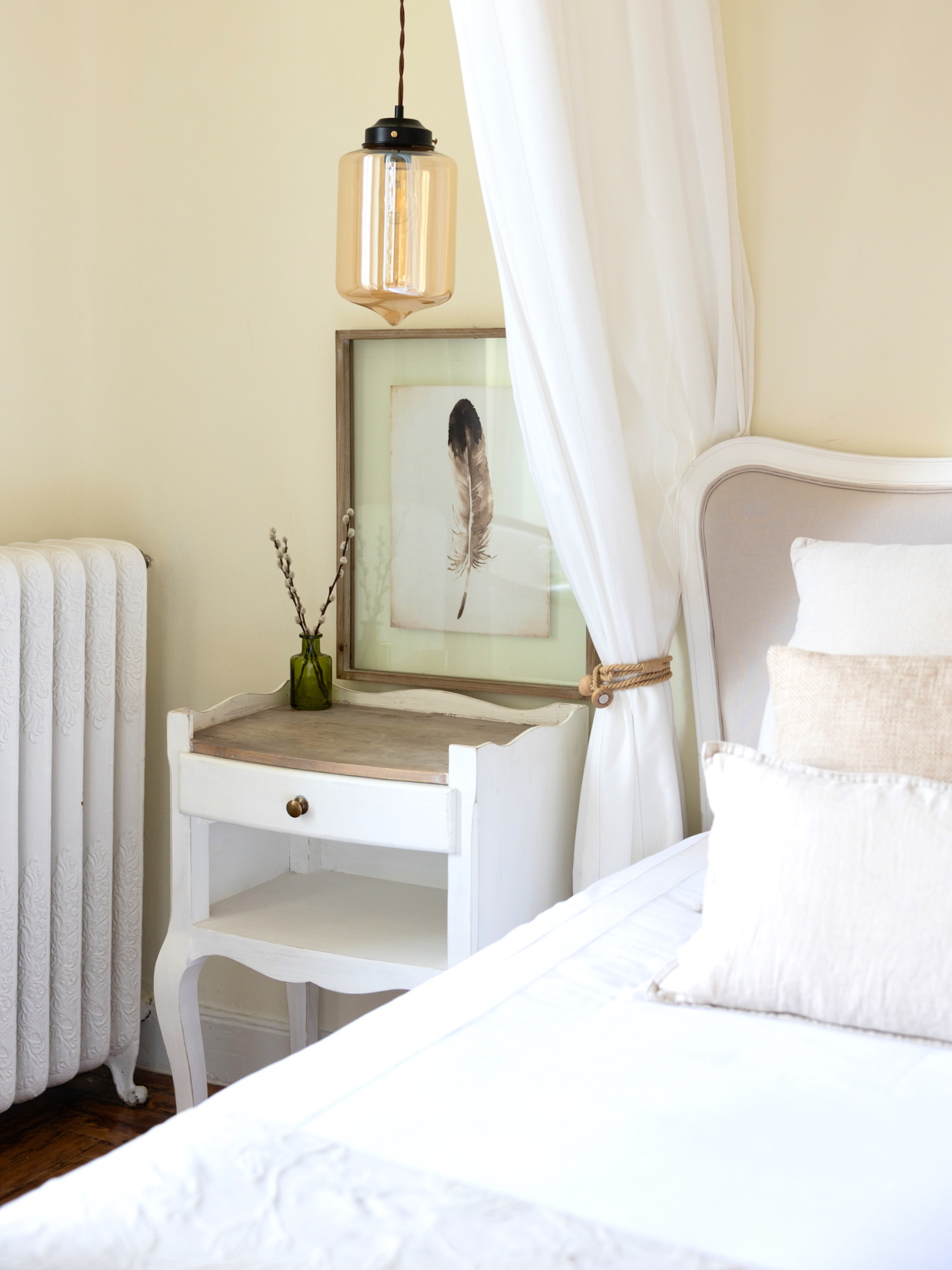 Dormitorio elegante en tonos blancos con mesita de noche vintage, lámpara colgante de cristal ámbar, cuadro de pluma y cortinas blancas con detalles en cuerda. Mesa auxiliar pintada en blanco con superficie de madera natural.