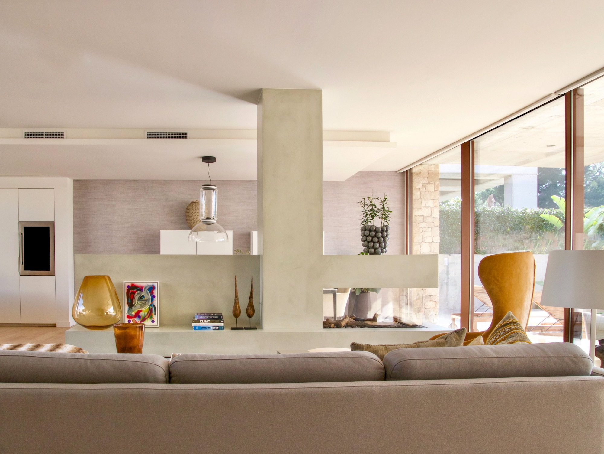 Sala de estar moderna y luminosa con chimenea decorativa central, sofá gris claro, ventanales amplios, sillón mostaza y elementos decorativos como jarrones y arte en tonos cálidos. Diseño minimalista con acabados en beige.
