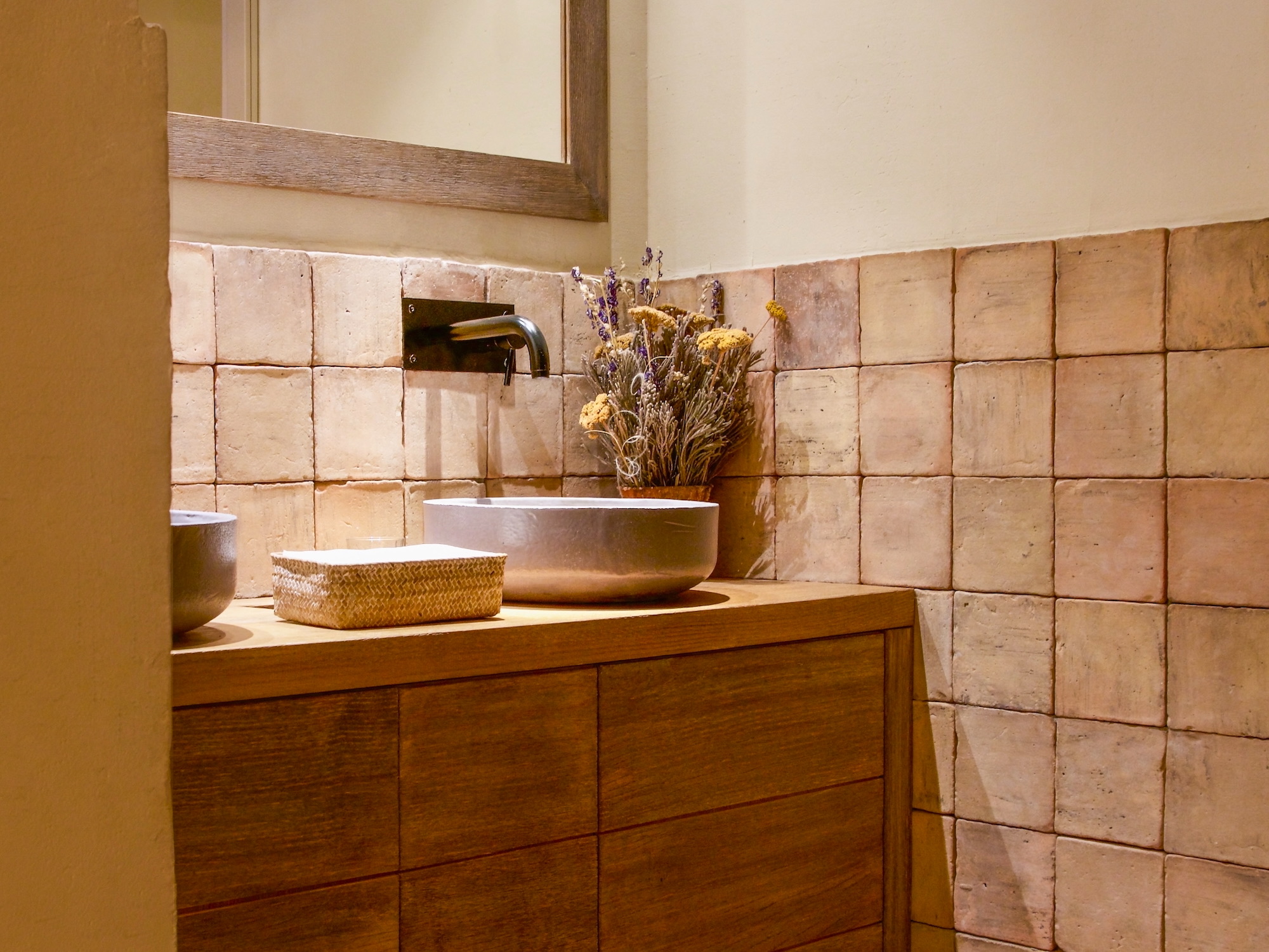 Baño de estilo rústico moderno con mueble de madera, lavabo sobre encimera blanco, azulejos terracota, espejo enmarcado en madera y arreglo floral seco decorativo