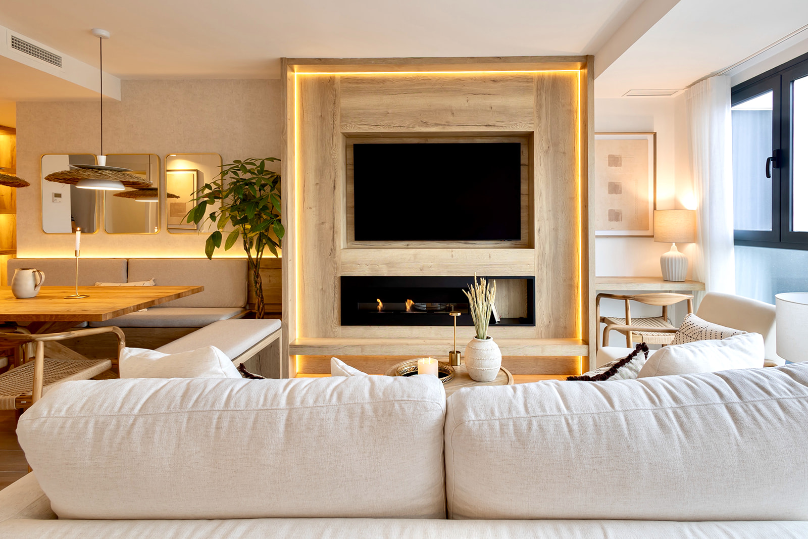 Sala de estar moderna con chimenea eléctrica y televisor enmarcados en madera con iluminación LED. Sofá blanco, mesa de comedor y decoración en tonos cálidos. Ambiente acogedor y luminoso.