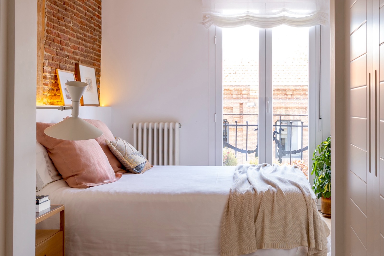 Dormitorio elegante con cama blanca, paredes de ladrillo, balcón y luz natural, decorado con cojines en tonos rosa y beige