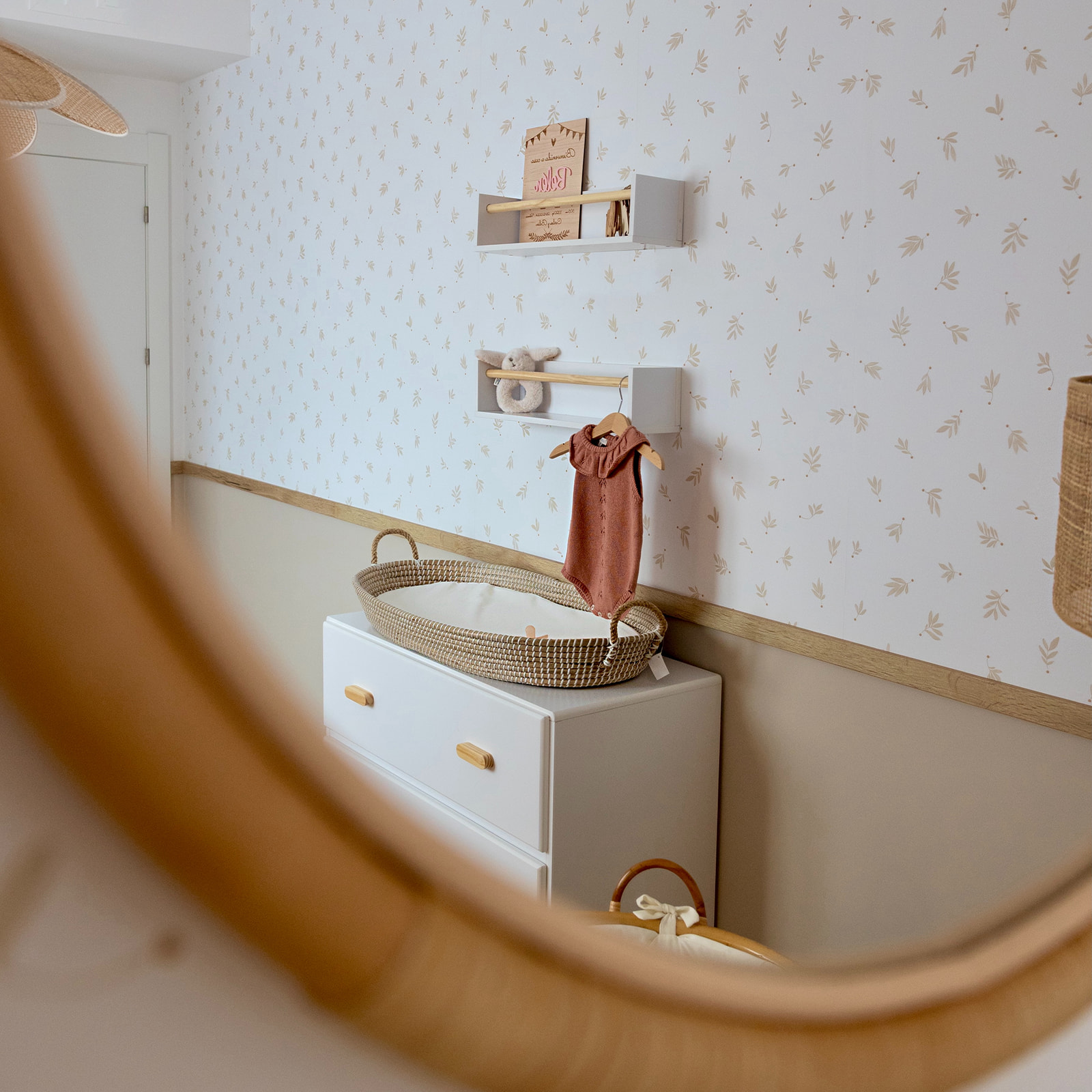 Habitación de bebé con estilo escandinavo, paredes con papel tapiz floral suave, cómoda blanca con cesta de mimbre, estantes flotantes y un espejo circular de madera en primer plano