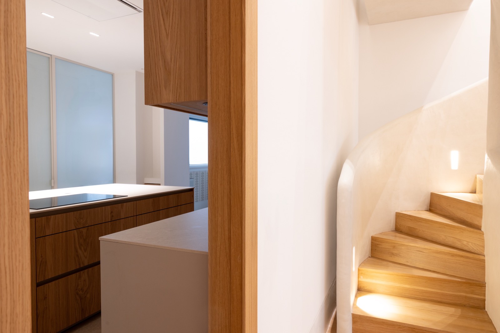 Escaleras de madera curvas con iluminación suave, junto a cocina minimalista con acabados de madera y superficies blancas