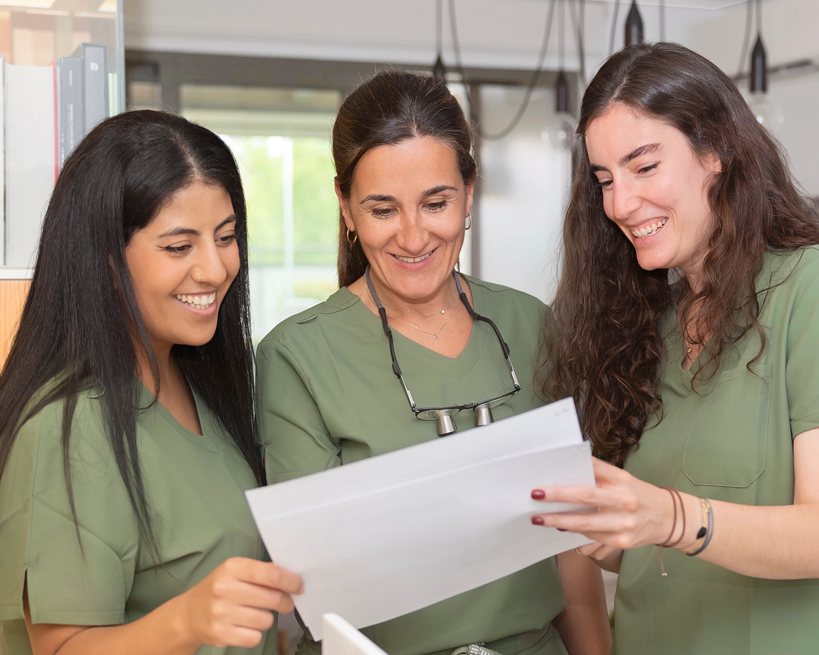 Tres profesionales de la salud en uniformes verdes sonriendo mientras revisan un documento juntas en un entorno médico. La imagen muestra un ambiente de colaboración y positividad en el trabajo.