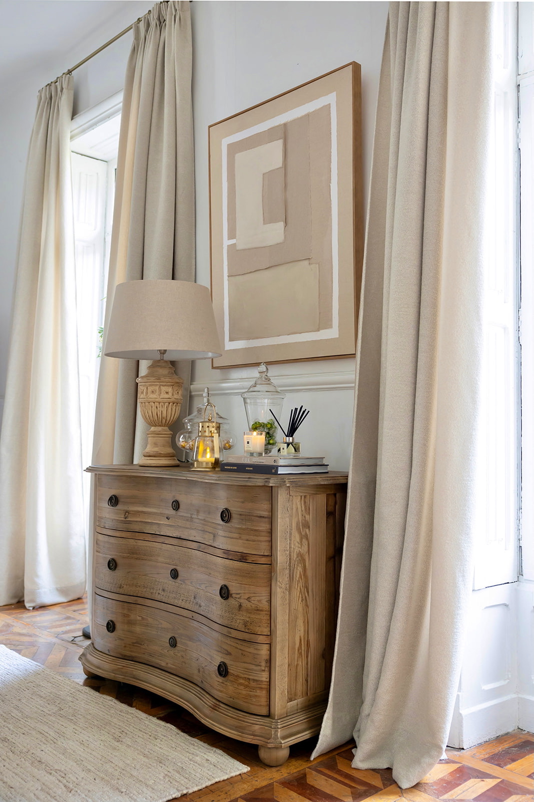 Cómoda de madera rústica con detalles curvos y tiradores negros, decorada con una lámpara vintage, velas y un cuadro abstracto en tonos beige. Cortinas claras y piso de parquet completan la escena elegante.
