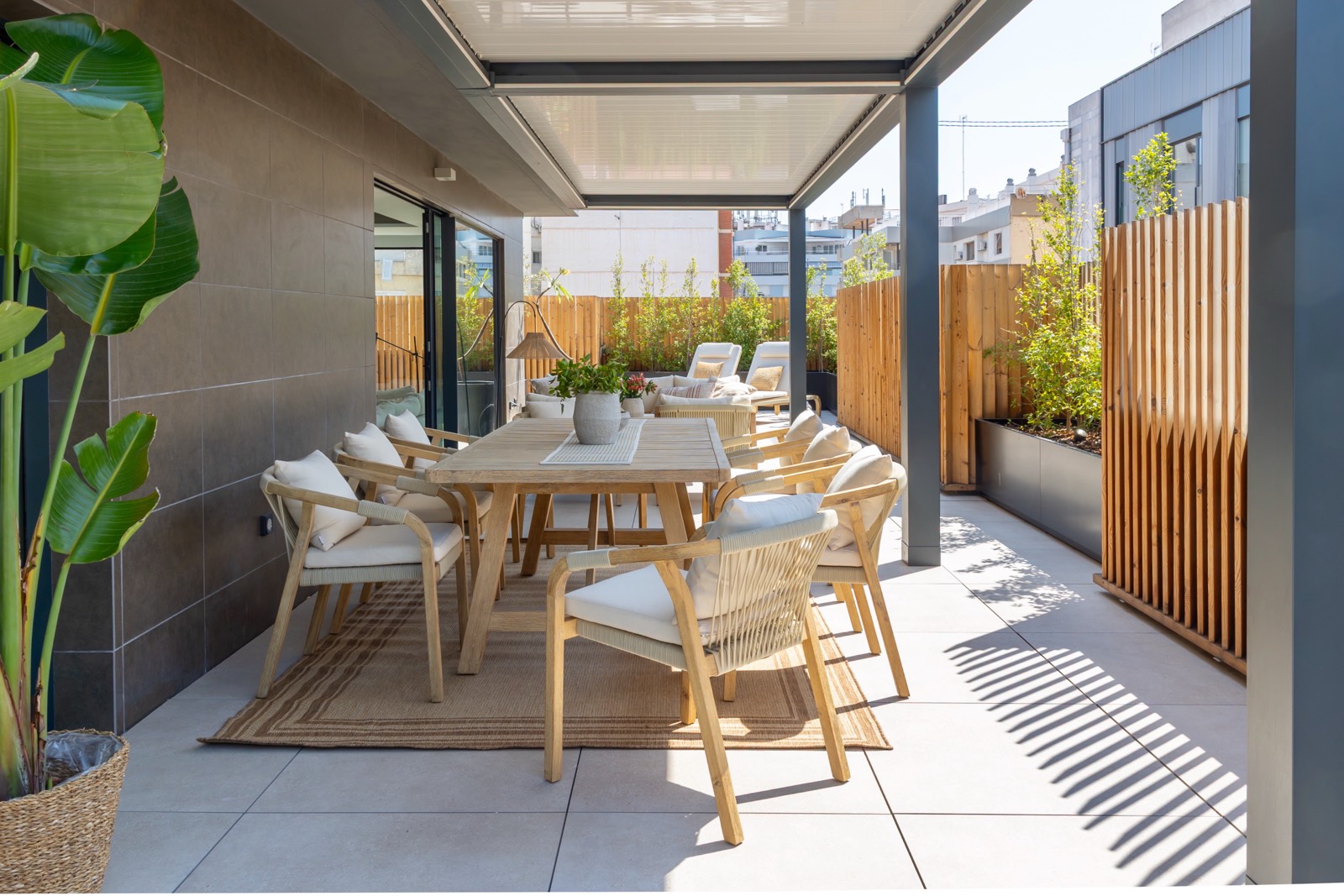 Terraza moderna con mesa de comedor de madera y sillas, rodeada de plantas y vallas de madera. Espacio exterior elegante y acogedor con techo retráctil y vista a edificios urbanos.