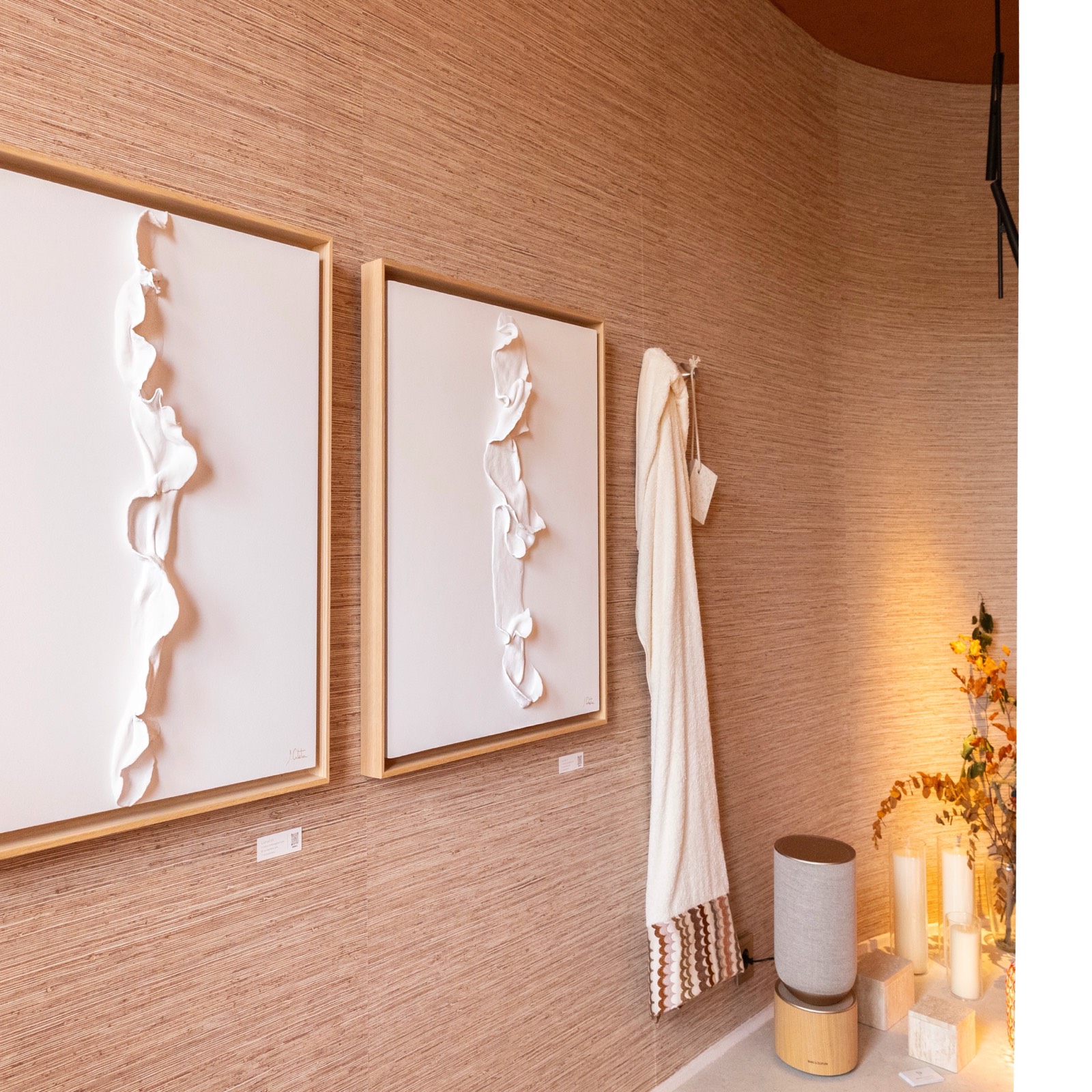 Dos cuadros de arte abstracto en blanco con textura en relieve, colgados junto a una toalla blanca y decoración minimalista con velas y florero