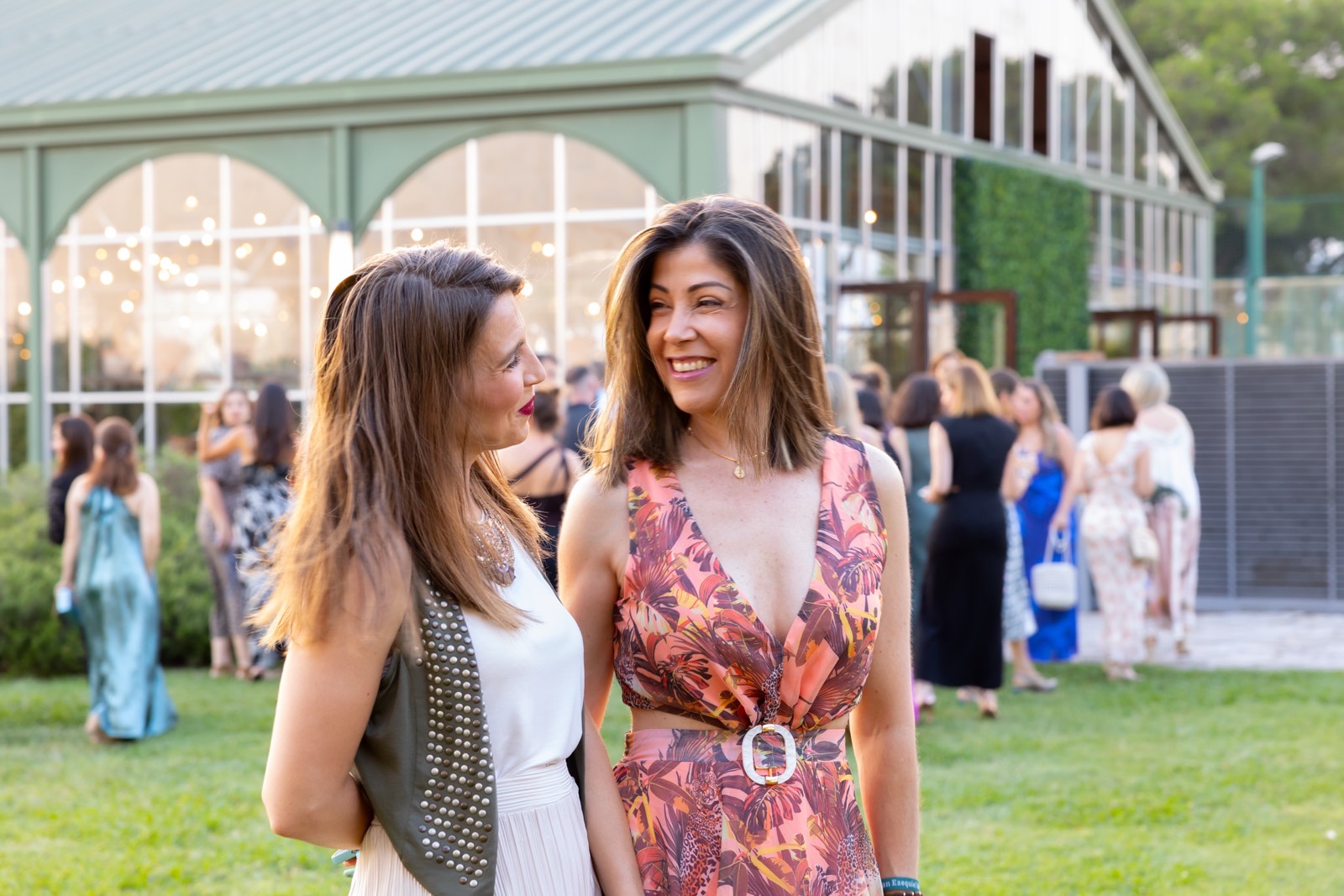 Dos mujeres sonrientes conversando en un evento al aire libre, con un edificio moderno y otras personas de fondo