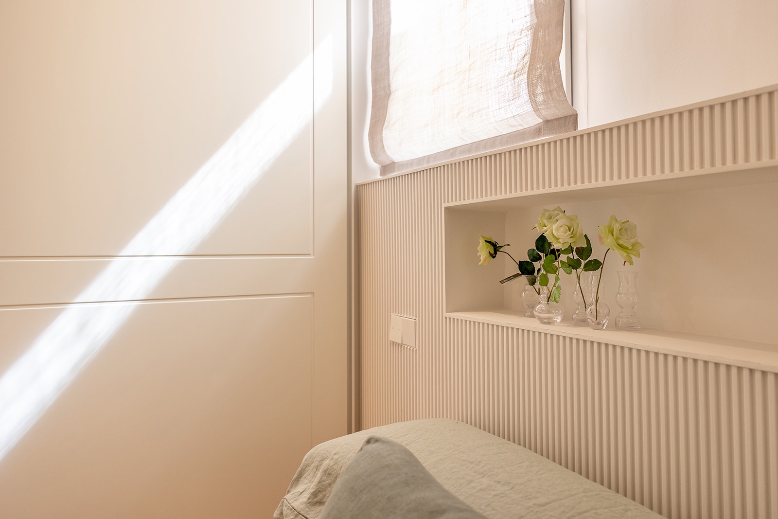 Habitación minimalista con paredes blancas y paneles acanalados. Una ventana con cortina blanca deja entrar luz natural. Un nicho en la pared contiene tres floreros con rosas blancas. Se ve parte de una cama con sábana gris claro.