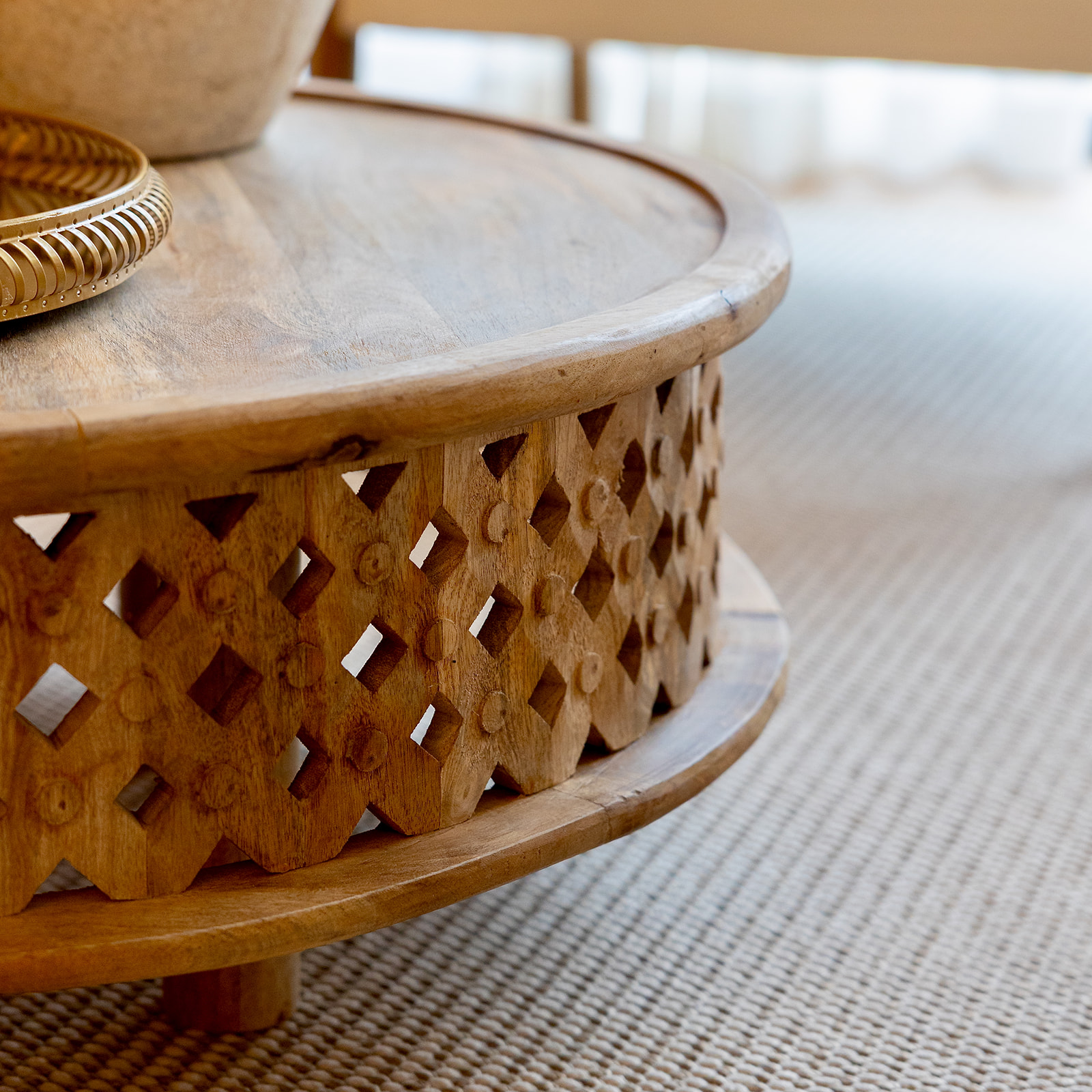 Mesa de centro circular de madera con diseño tallado en el lateral. La mesa tiene un patrón de diamantes calados y está colocada sobre una alfombra de color claro. En la superficie de la mesa se ve parcialmente un cuenco de mimbre.
