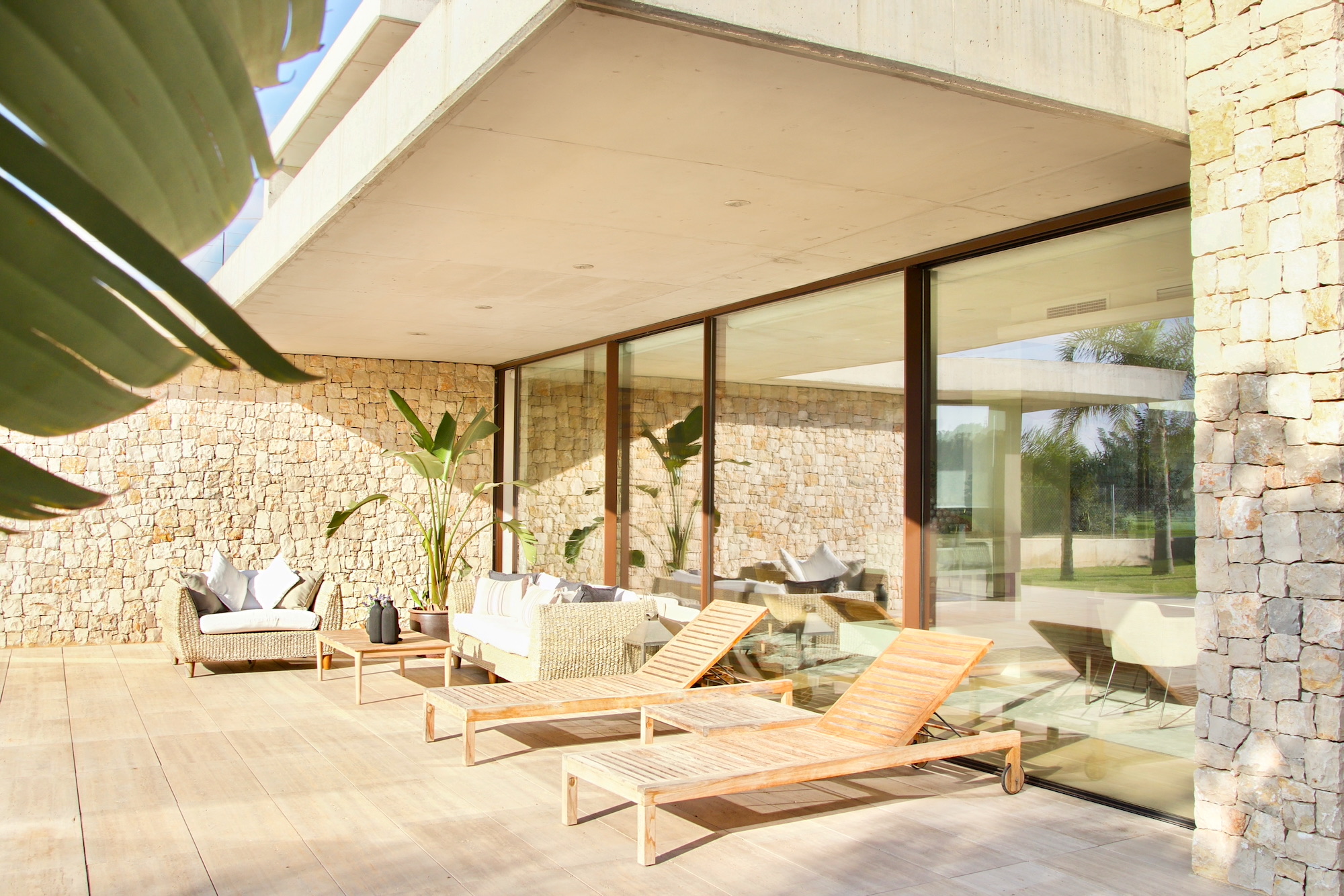 Elegante terraza exterior con paredes de piedra natural, grandes ventanales y muebles de exterior que incluyen tumbonas de madera y sofás de ratán. Decorada con plantas tropicales y acabados modernos.