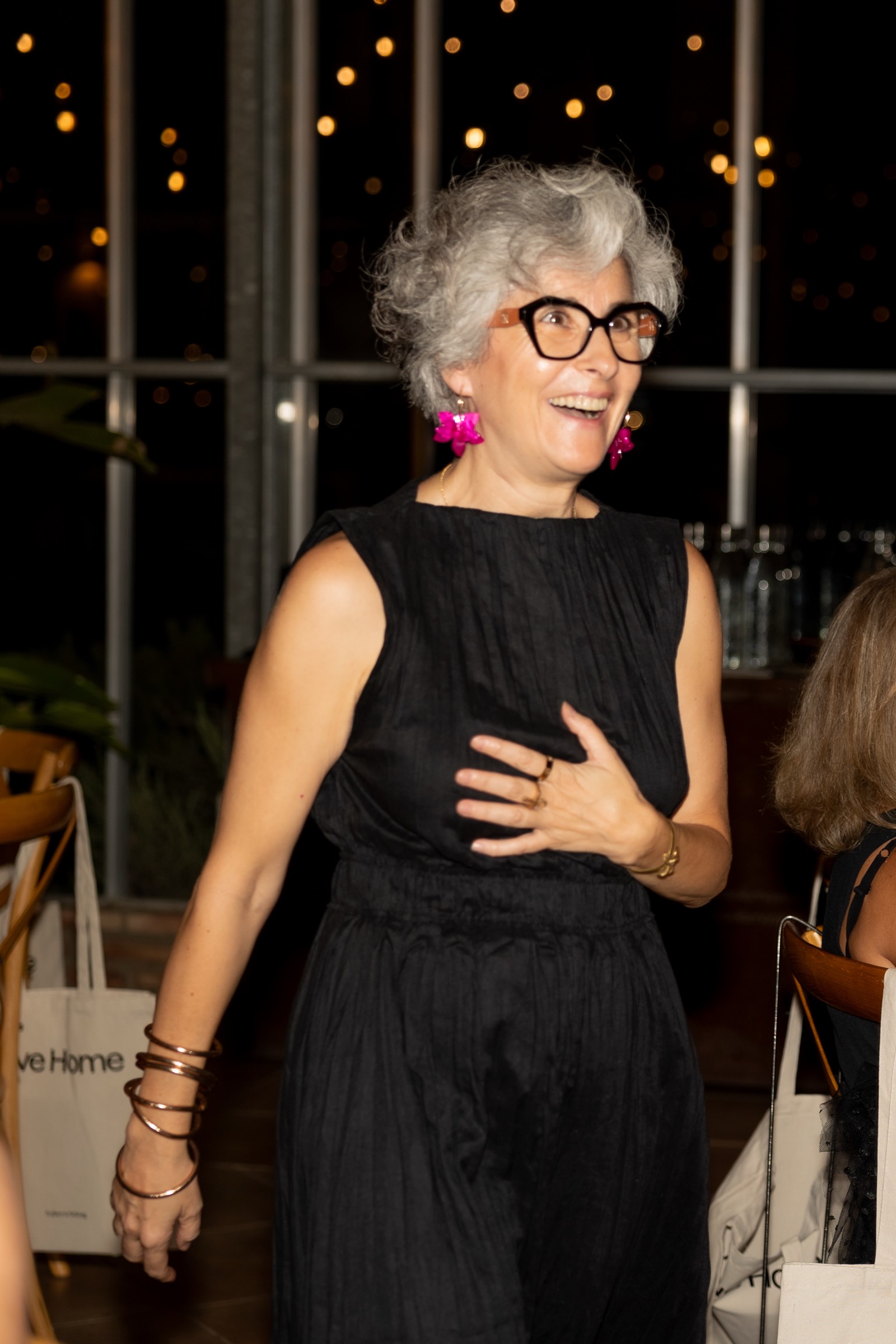 Señora mayor sonriente con cabello gris rizado, gafas y aretes rosados, vistiendo un vestido negro en un evento nocturno