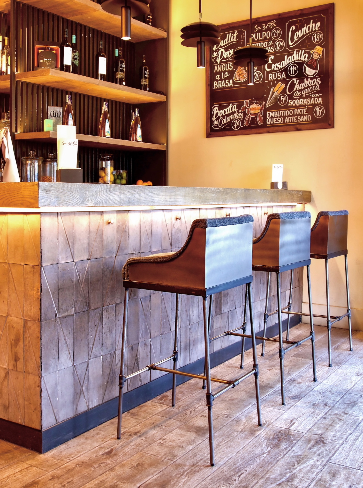 Interior de un bar de tapas con barra metálica decorativa, taburetes altos tapizados en cuero, estanterías de madera con botellas y un menú de pizarra en la pared que muestra diferentes platos españoles y sus precios