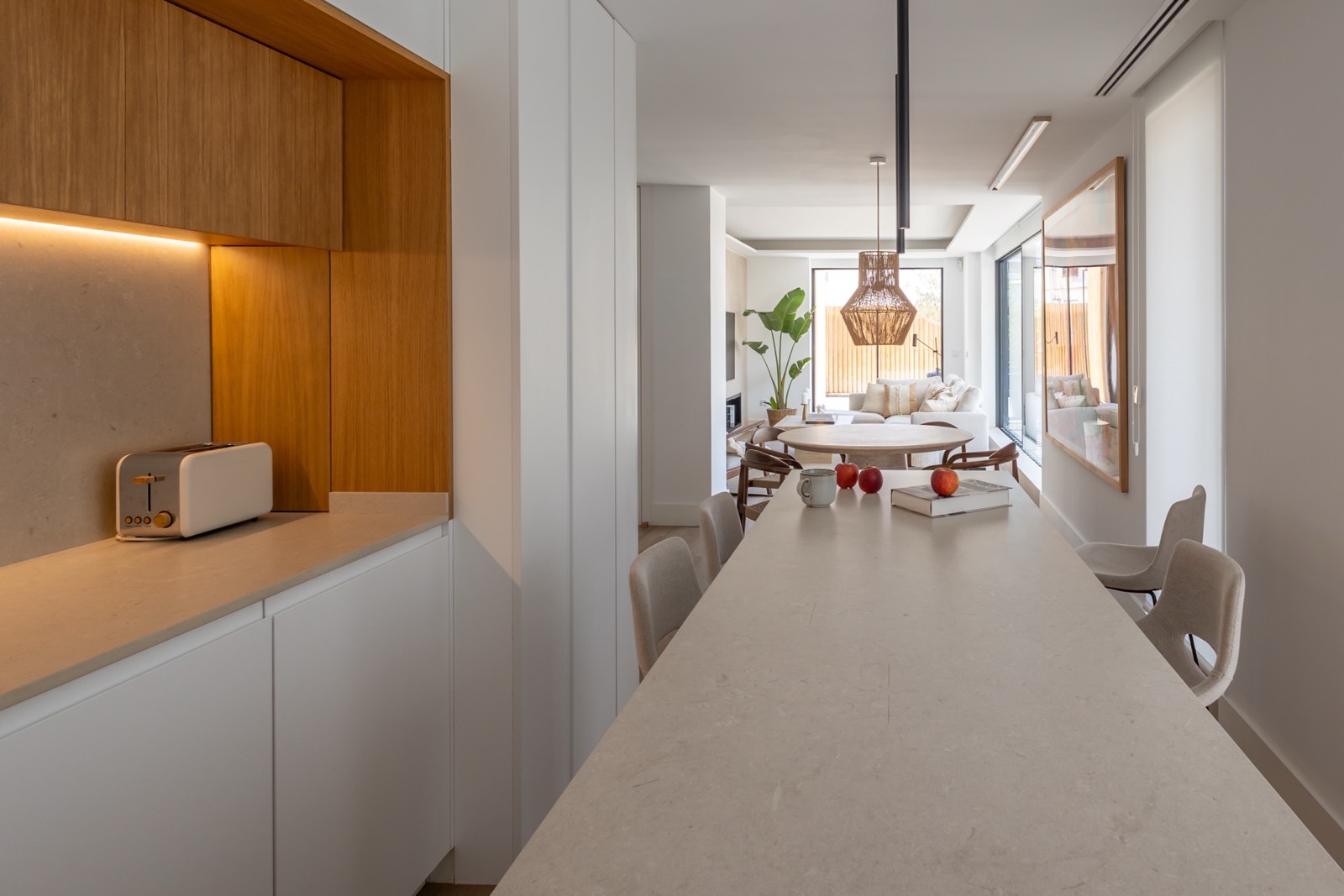 Cocina moderna abierta con isla de mármol blanco, armarios de madera y blancos, que se conecta con una sala de estar luminosa. Decoración minimalista con toques cálidos y naturales.