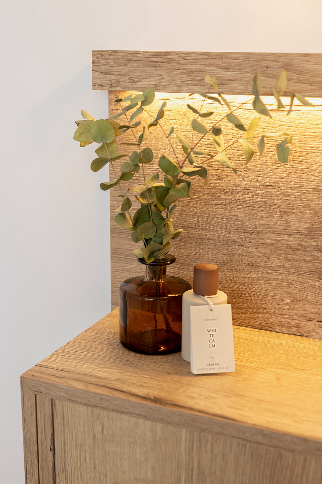 Estante de madera con iluminación cálida, florero de vidrio marrón con hojas verdes y un frasco blanco de perfume sobre una superficie de madera clara. Diseño minimalista y acogedor.