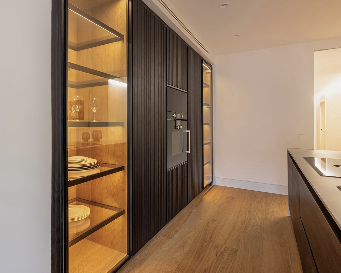 Cocina moderna con estantería empotrada iluminada mostrando vajilla y copas. Muebles de madera oscura con diseño acanalado. Electrodomésticos integrados y suelo de madera clara.