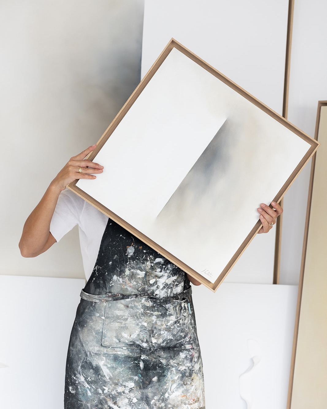 Artista con delantal manchado de pintura sosteniendo un lienzo en blanco enmarcado frente a su rostro en un estudio de arte minimalista