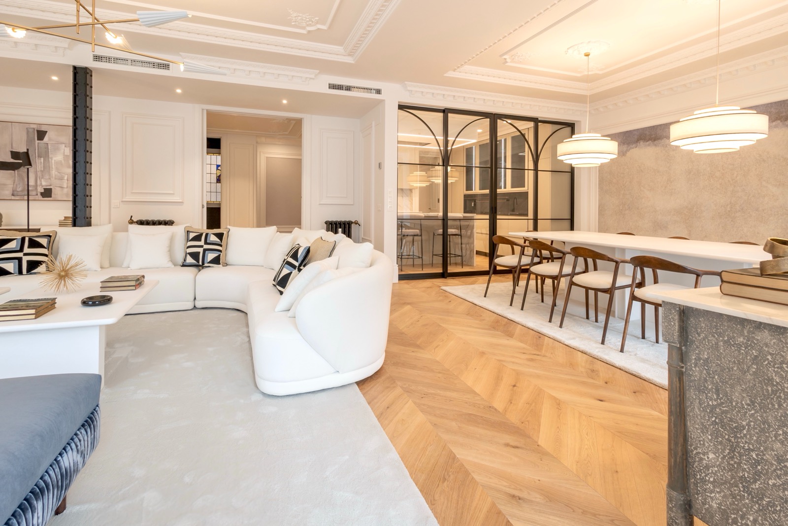 Amplia sala de estar y comedor de estilo moderno y elegante con sofá blanco, mesa de comedor, sillas de madera, lámparas colgantes y puertas de cristal que separan la cocina. El espacio tiene techos altos con molduras y suelo de parquet.