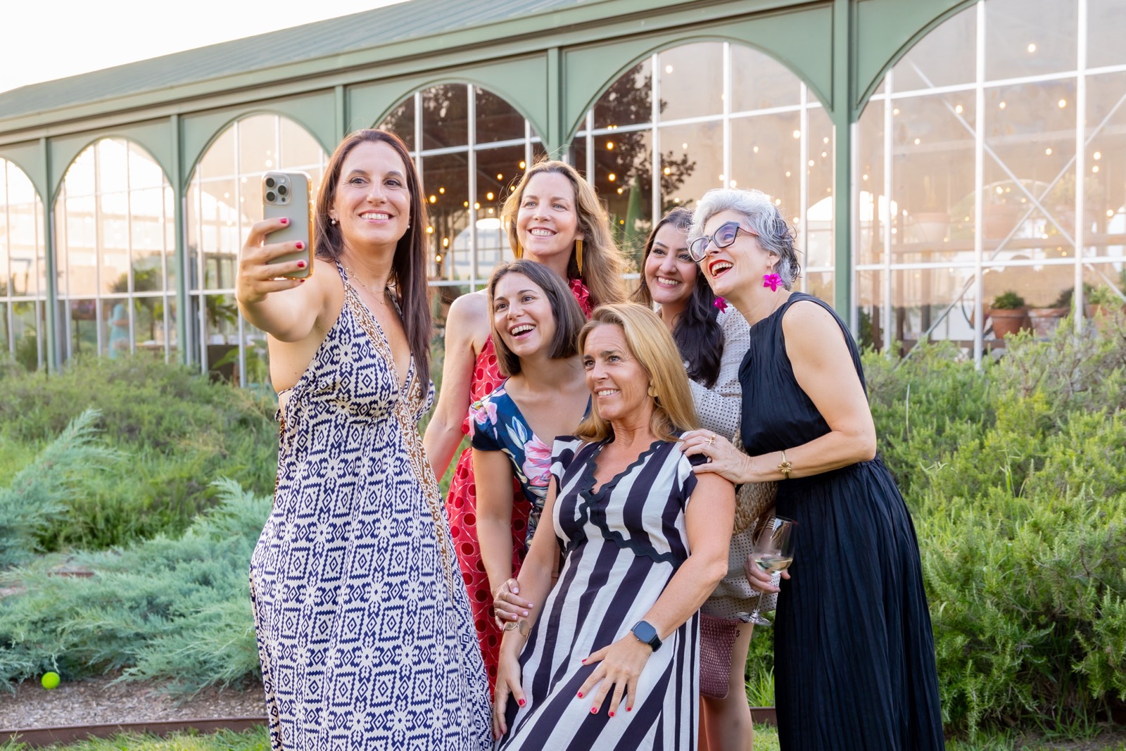 Grupo de mujeres de diferentes edades sonriendo juntas, tomándose un selfie frente a un edificio con ventanales y jardines