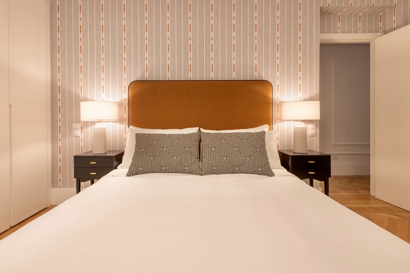 Dormitorio moderno y elegante con cabecero marrón, papel tapiz a rayas, lámparas de mesa blancas y cama con sábanas blancas y cojines estampados. El suelo es de parquet y hay una puerta abierta visible.