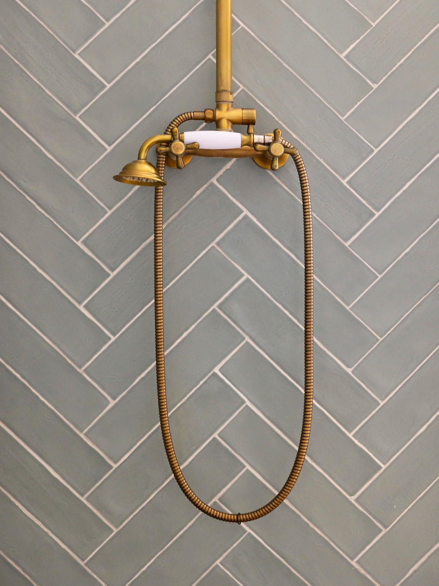 Ducha de estilo vintage en latón dorado con manguera flexible sobre pared de azulejos grises en patrón espiga. La regadera cuenta con acabados clásicos y controles cruzados tradicionales.