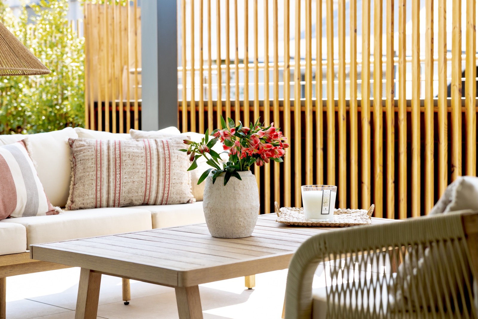 Terraza acogedora con sofá blanco, cojines a rayas, mesa de café de madera, florero con flores rojas y división de bambú. Ambiente luminoso y relajante con estilo bohemio.