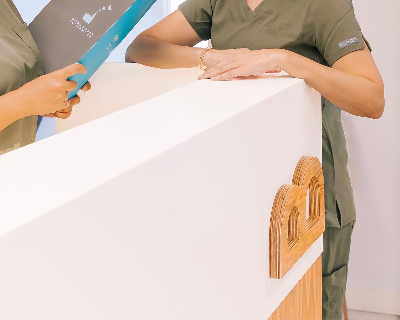 Recepcionista en uniforme verde atendiendo a un paciente en el mostrador de una clínica dental. Se ve una sierra dental en mano de otra persona y un logotipo de madera en el mostrador.