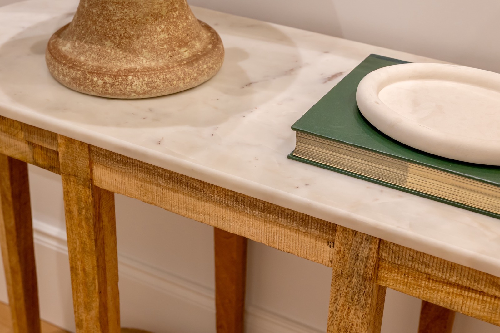 Mesa de madera con superficie de mármol blanco, sobre la que reposan una base de lámpara dorada, un libro verde y un plato blanco. La mesa tiene patas de madera y está colocada contra una pared blanca.